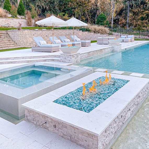 Outdoor pool and hot tub with white glass mosaic tile, firepit with beige stacked limestone surround and blue rocks, and white paver pool deck.